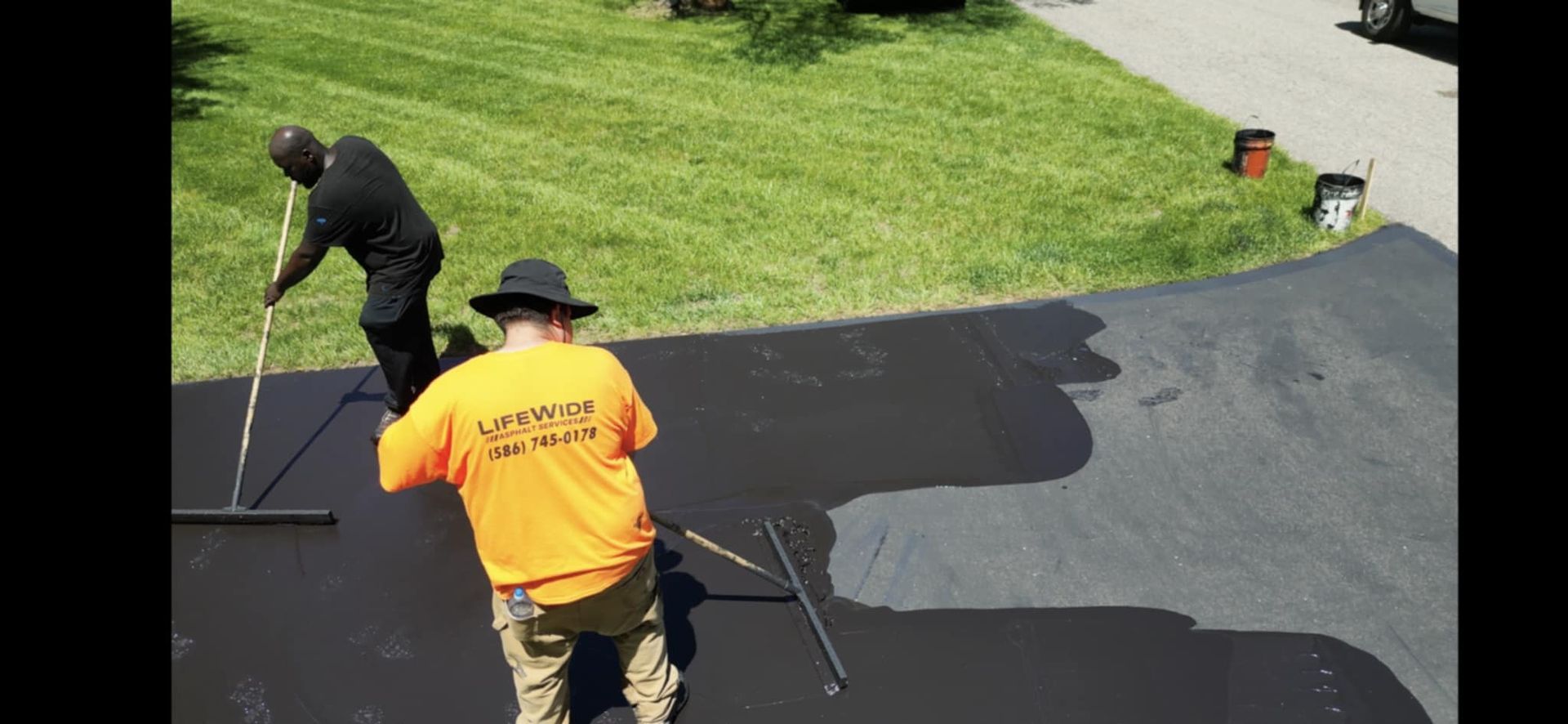 Two people sealcoating a dark driveway. One is spreading with a long handle brush. Green grass background.