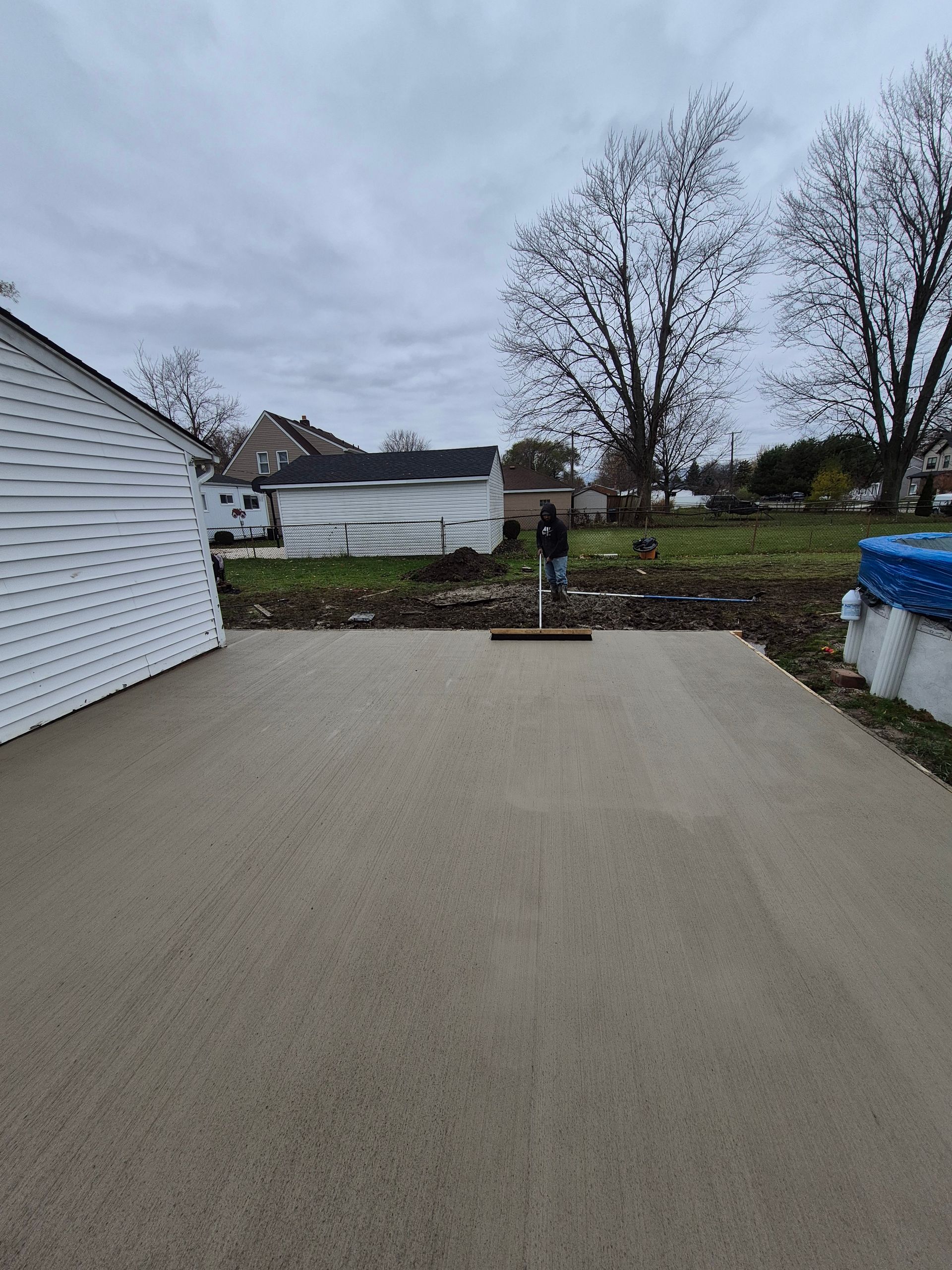 Person raking a newly poured concrete patio in a backyard under a cloudy sky.