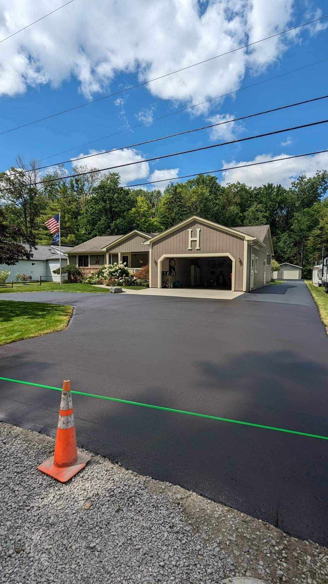 Newly paved asphalt driveway leading to a house with a garage; orange traffic cone in foreground.