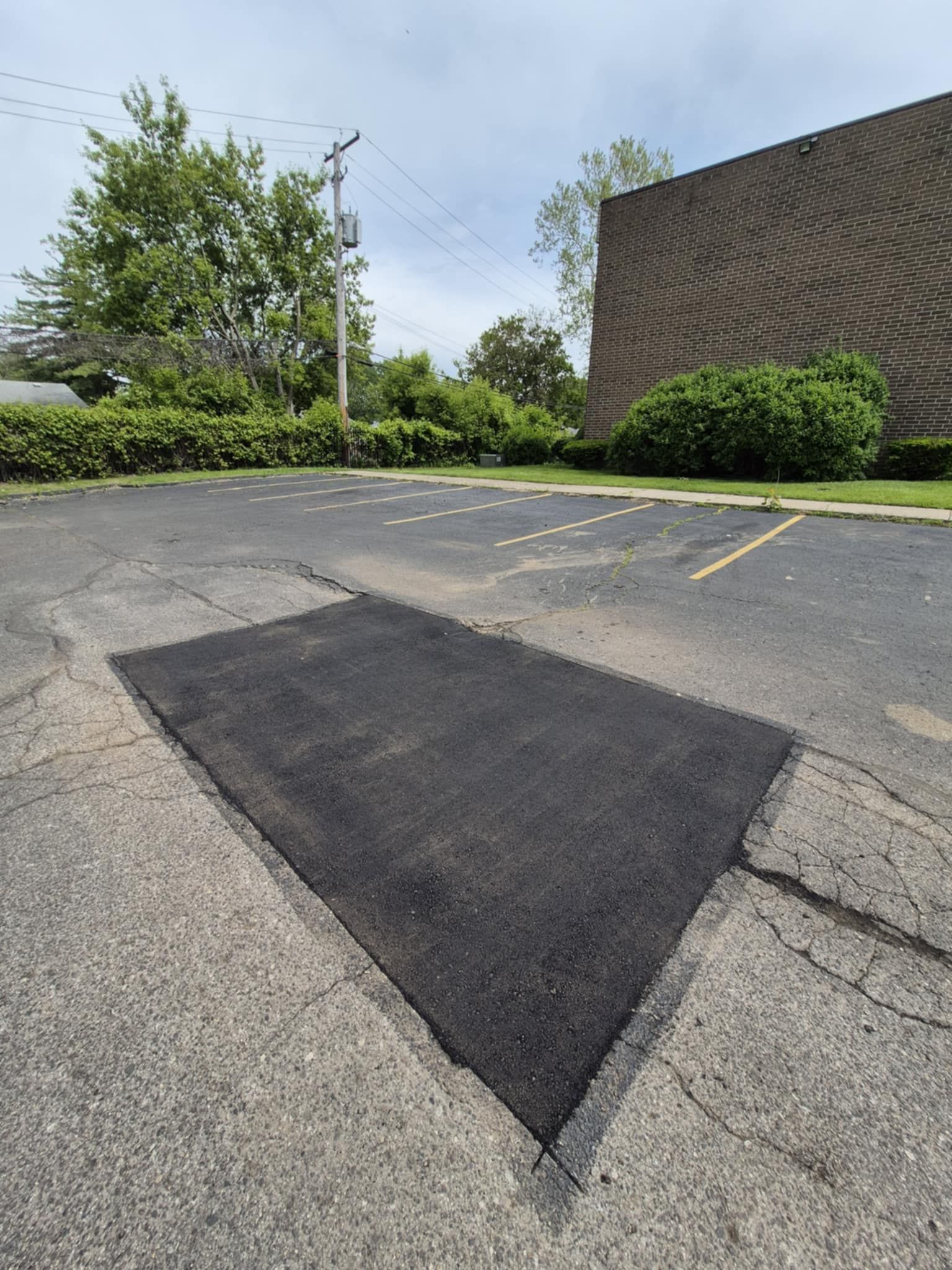 Asphalt parking lot with new, dark patch; trees and brick building in background.