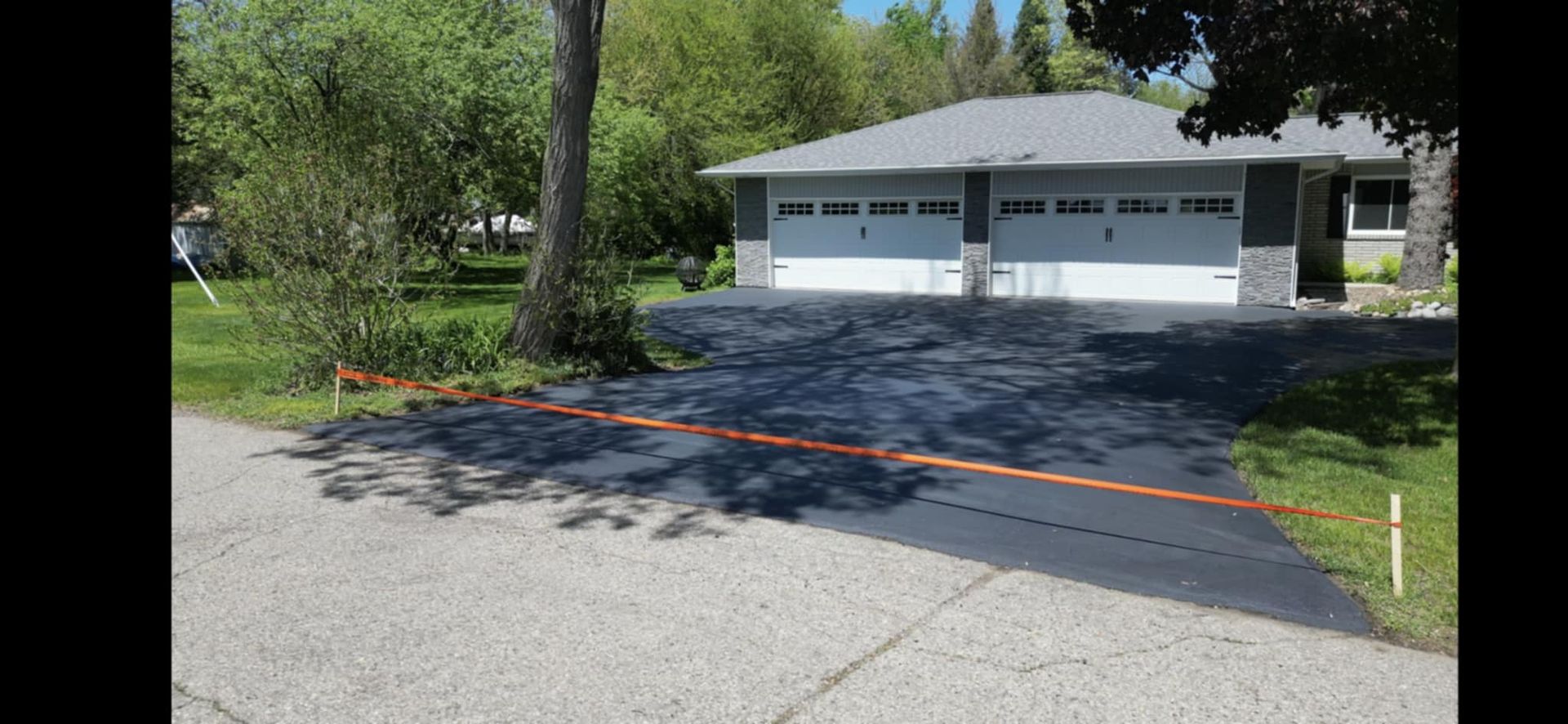 A house with a black driveway and two garage doors. Trees and green grass are around the house.