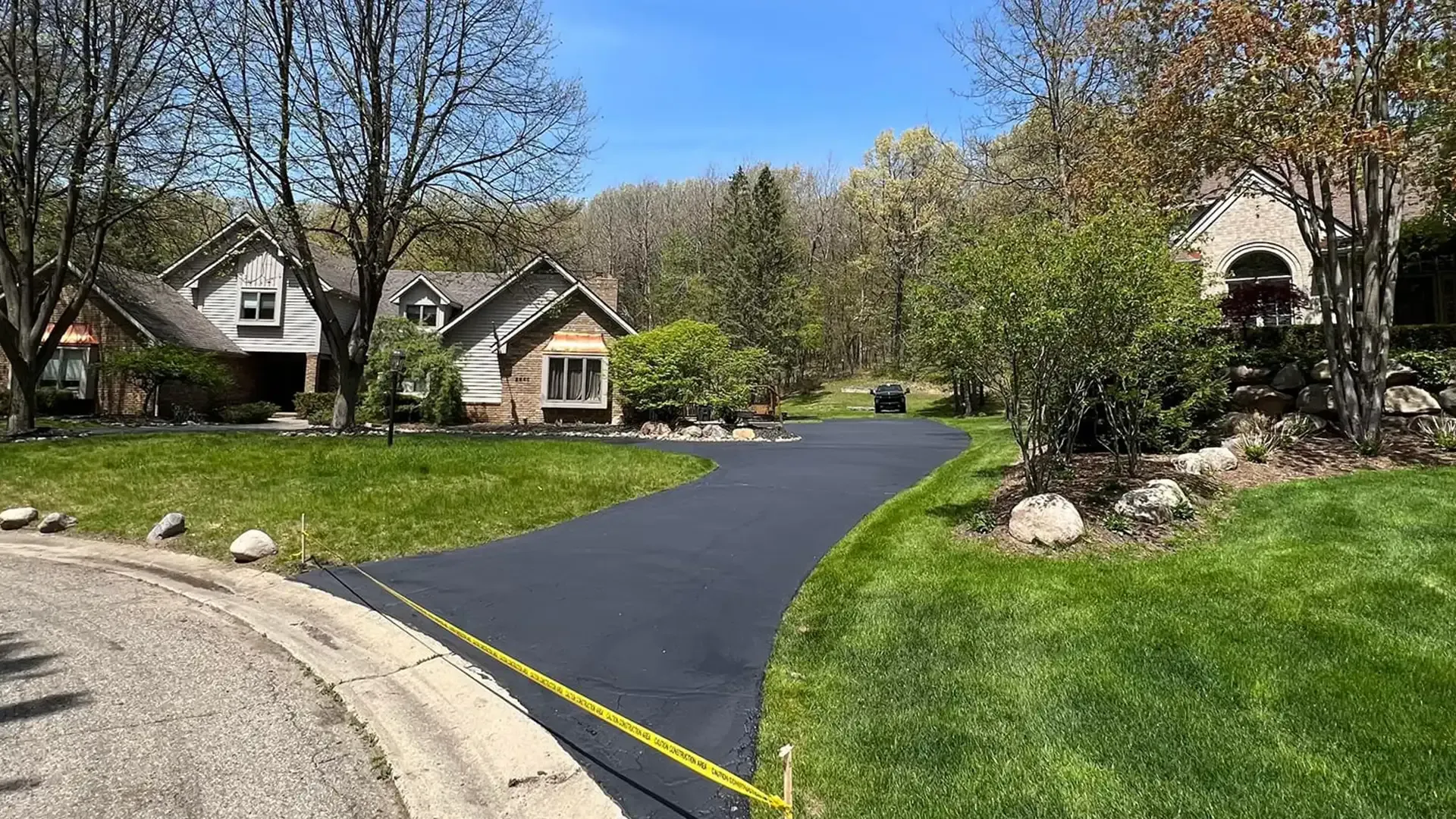Paved driveway leading to two houses with lush green lawns and trees against a blue sky.