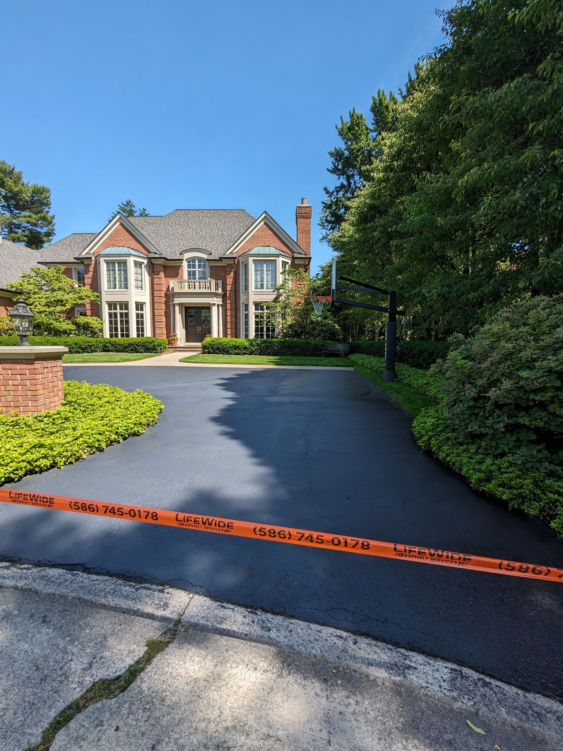 Newly paved black asphalt driveway leading to a large brick house. Orange caution tape stretches across the drive.