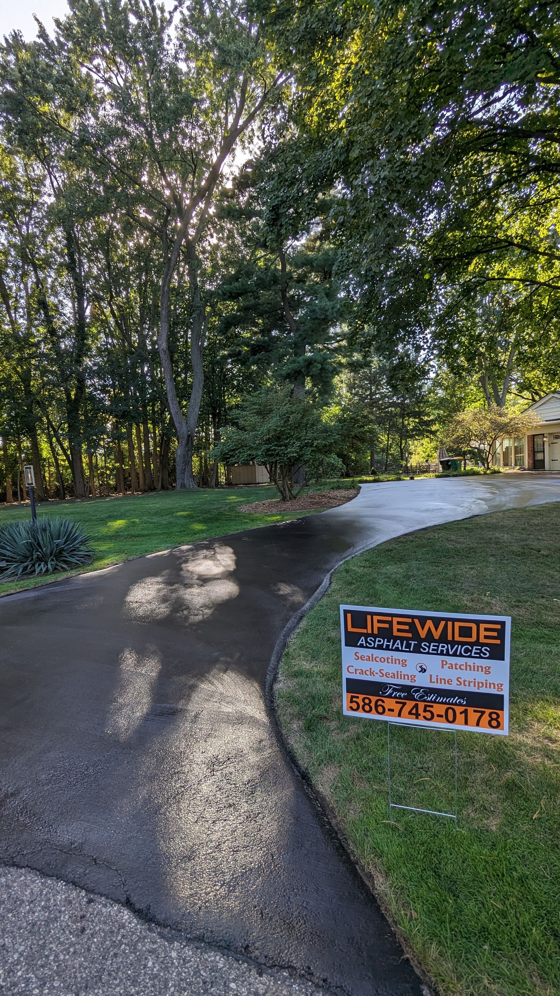 A sign advertising Lifewide Asphalt Sealcoating on a driveway, with trees and a house in the background.