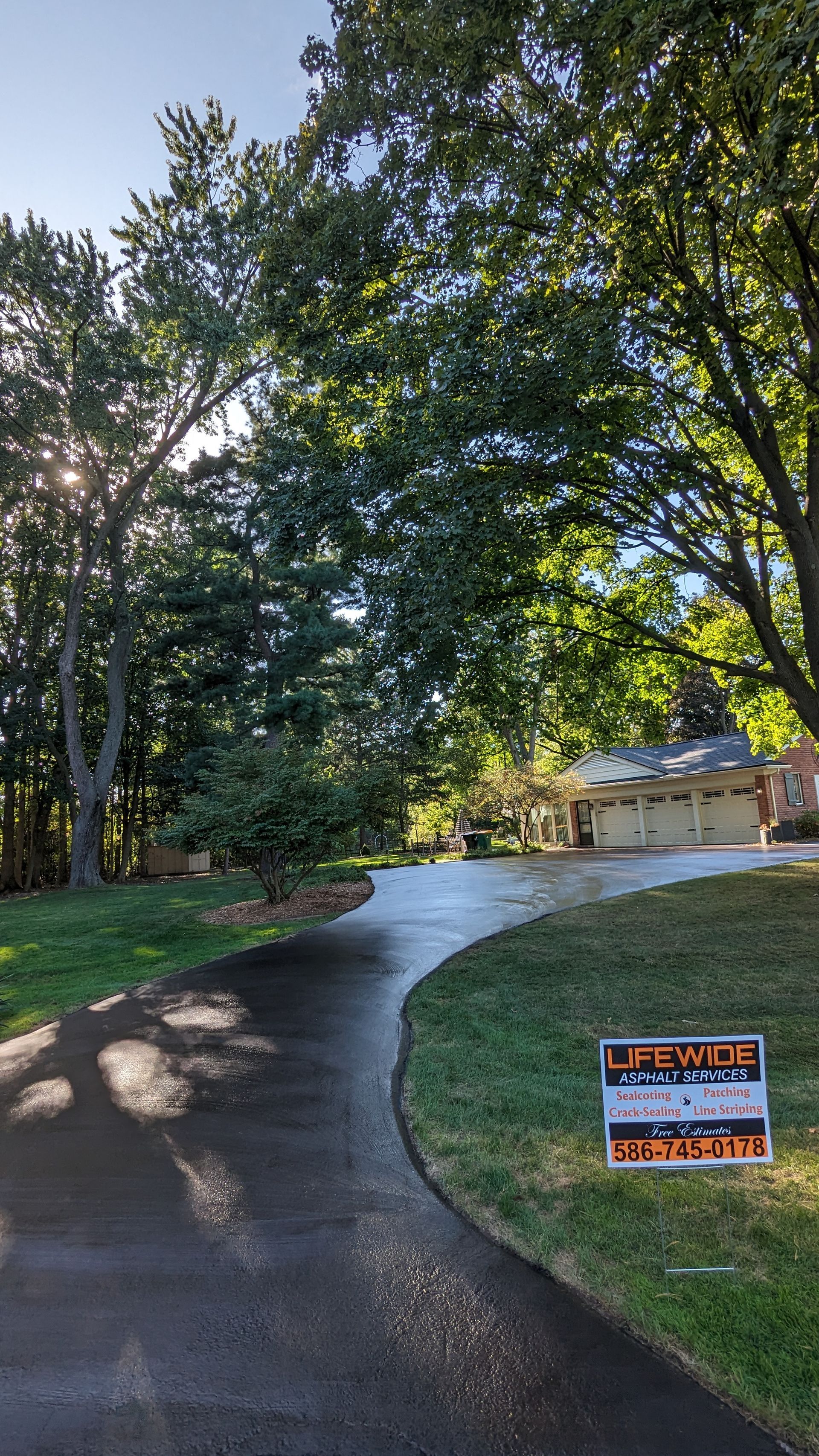 Asphalt driveway leading to a house, flanked by green grass and trees; a sign is on the right.