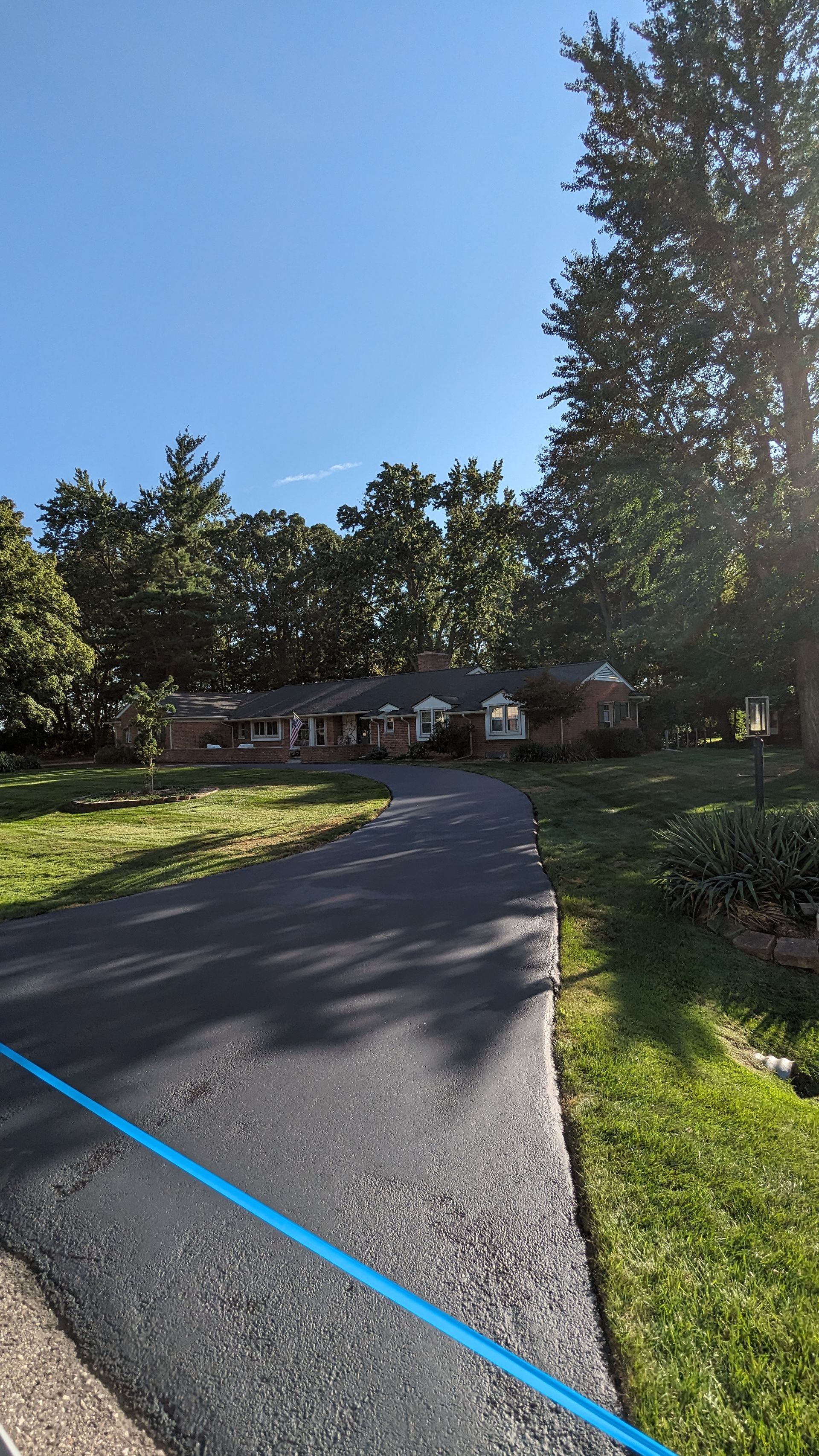 Asphalt driveway leading to a one-story house surrounded by trees under a blue sky.