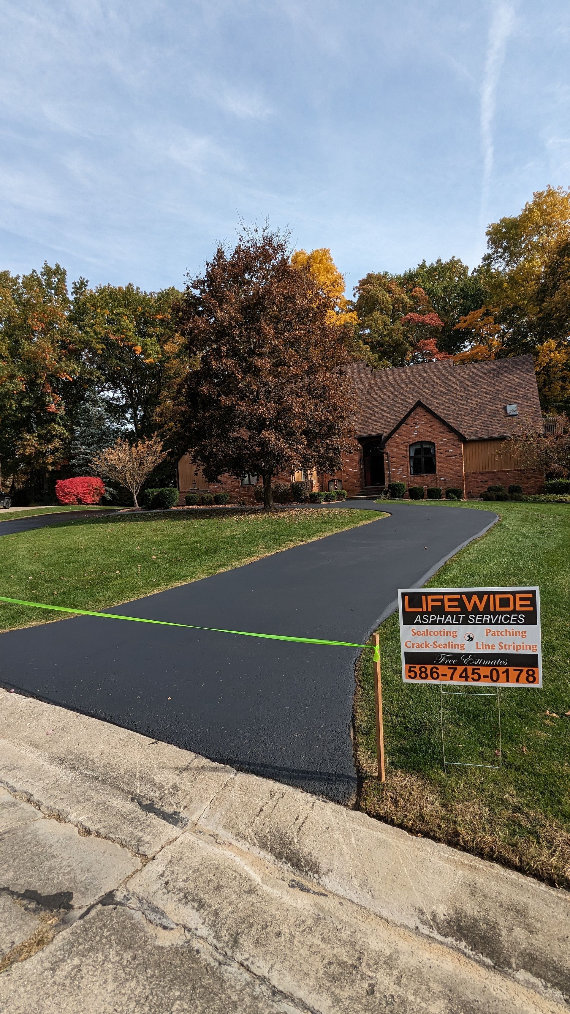 Newly paved asphalt driveway leading to a brick house on a sunny day with fall foliage.