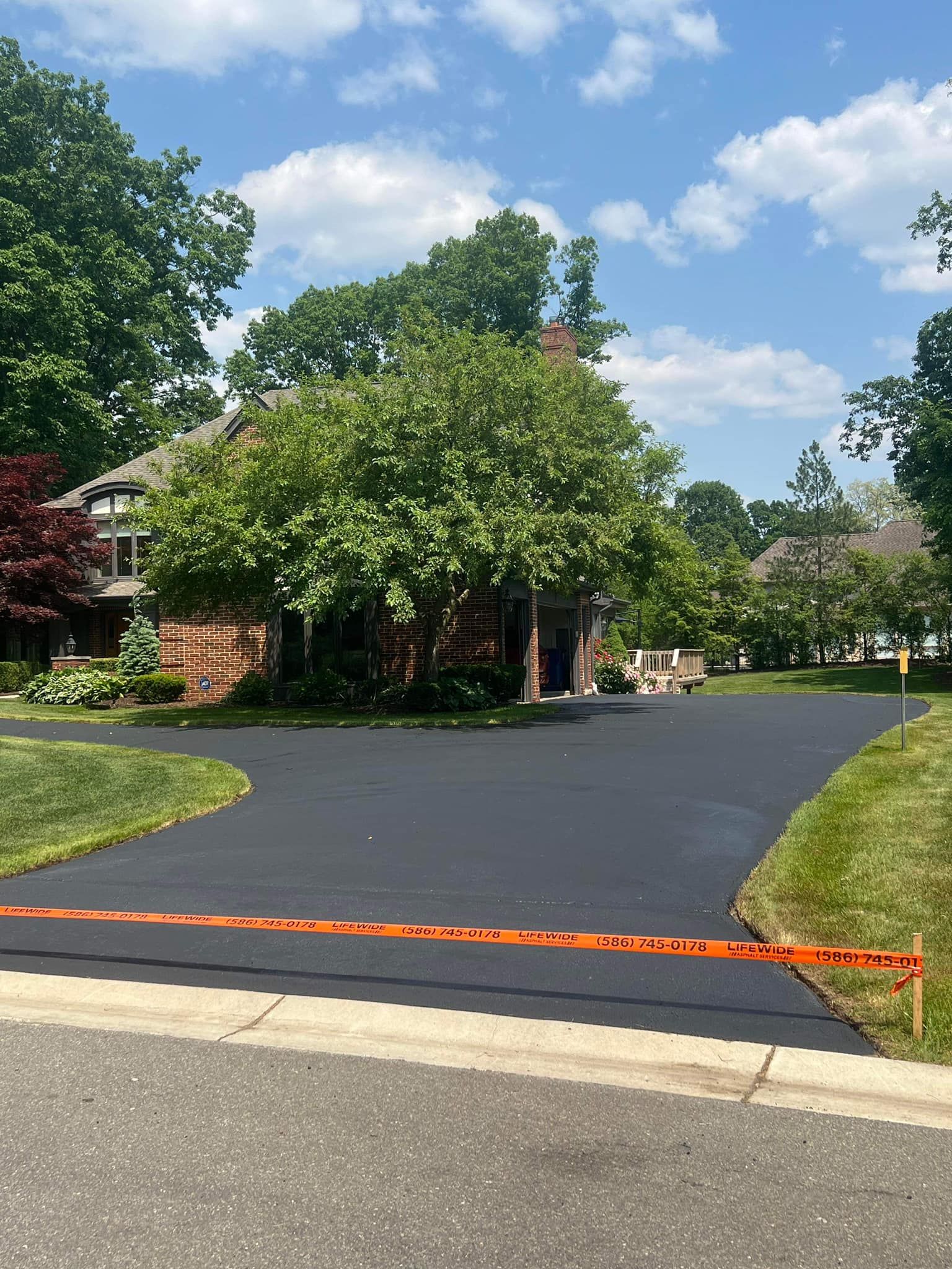 Newly paved black asphalt driveway in front of a brick house with green trees and blue sky. Orange tape across driveway.