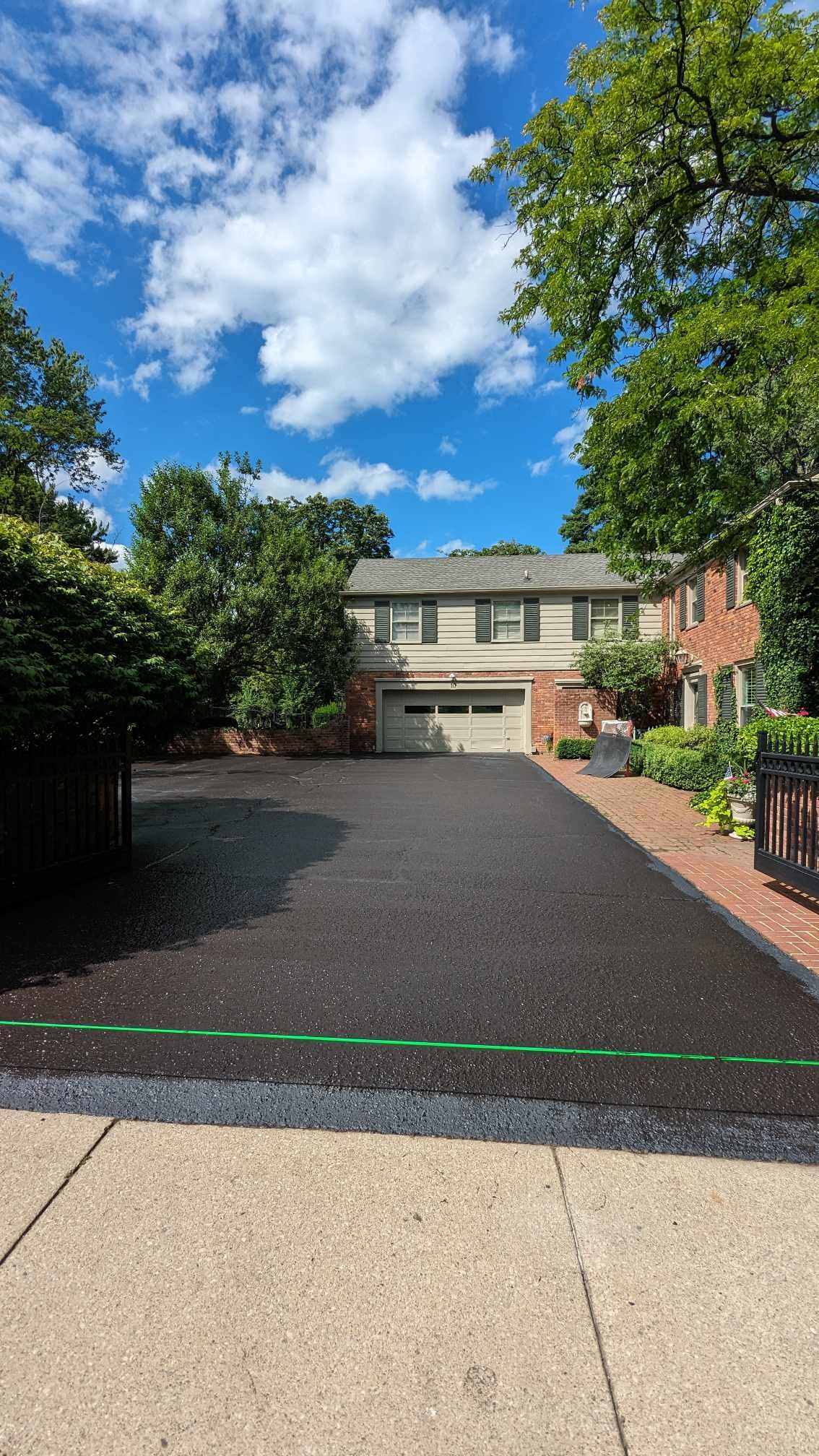 Asphalt driveway leading to a two-story house with a garage, trees, and blue sky.