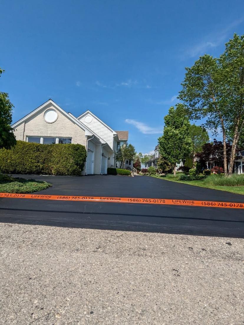 Newly paved black asphalt driveway with orange safety tape in front of a light-colored house under a bright blue sky.