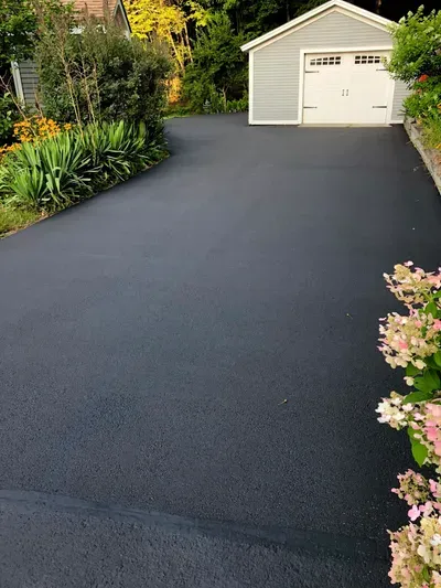 Freshly sealed black asphalt driveway leading to a light gray garage with white doors, surrounded by greenery.