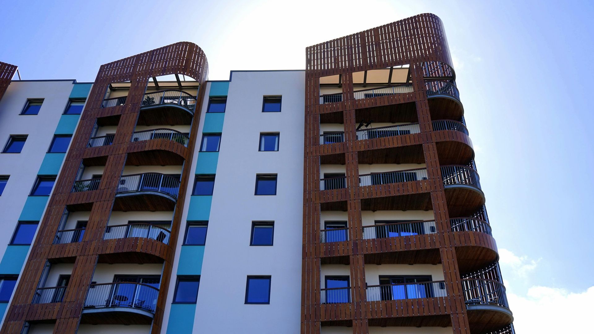 Multi-story apartment building with balconies; teal, white, and brown facade against a bright blue sky.