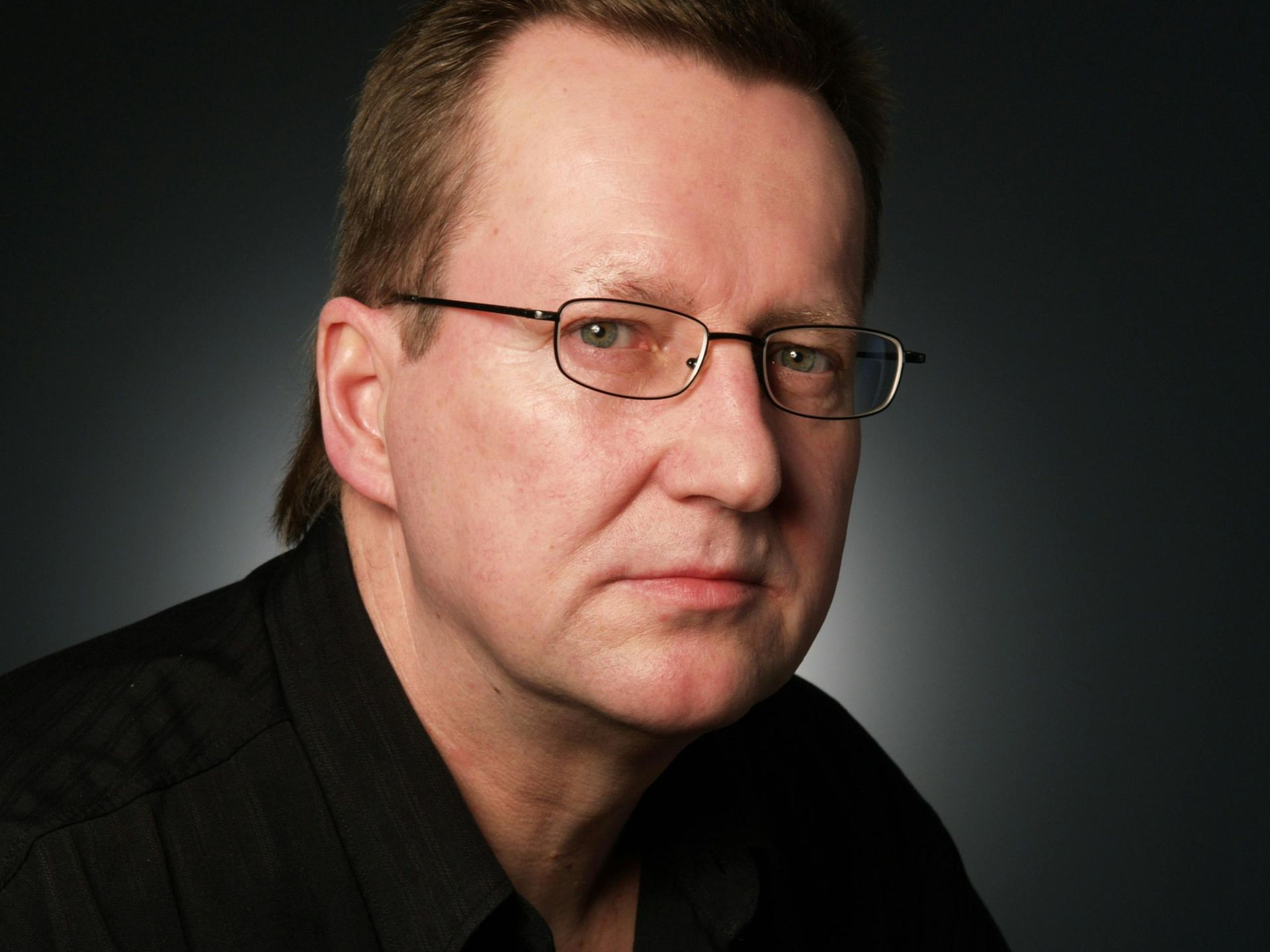 Middle-aged man wearing glasses, looking serious, wearing a black shirt, against a dark gray background.