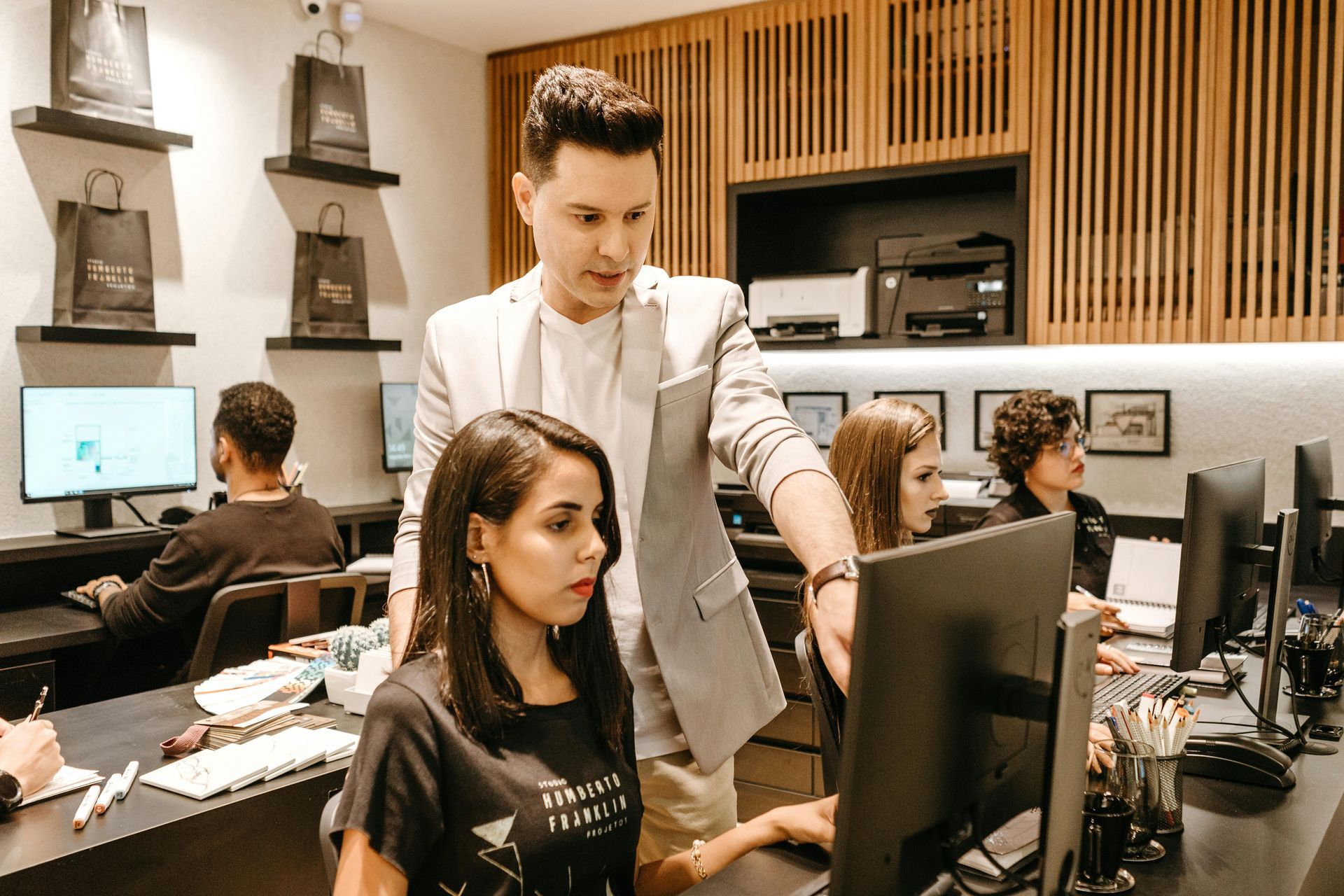 A man points at a computer screen, instructing a woman. They sit in a modern office with other workers.