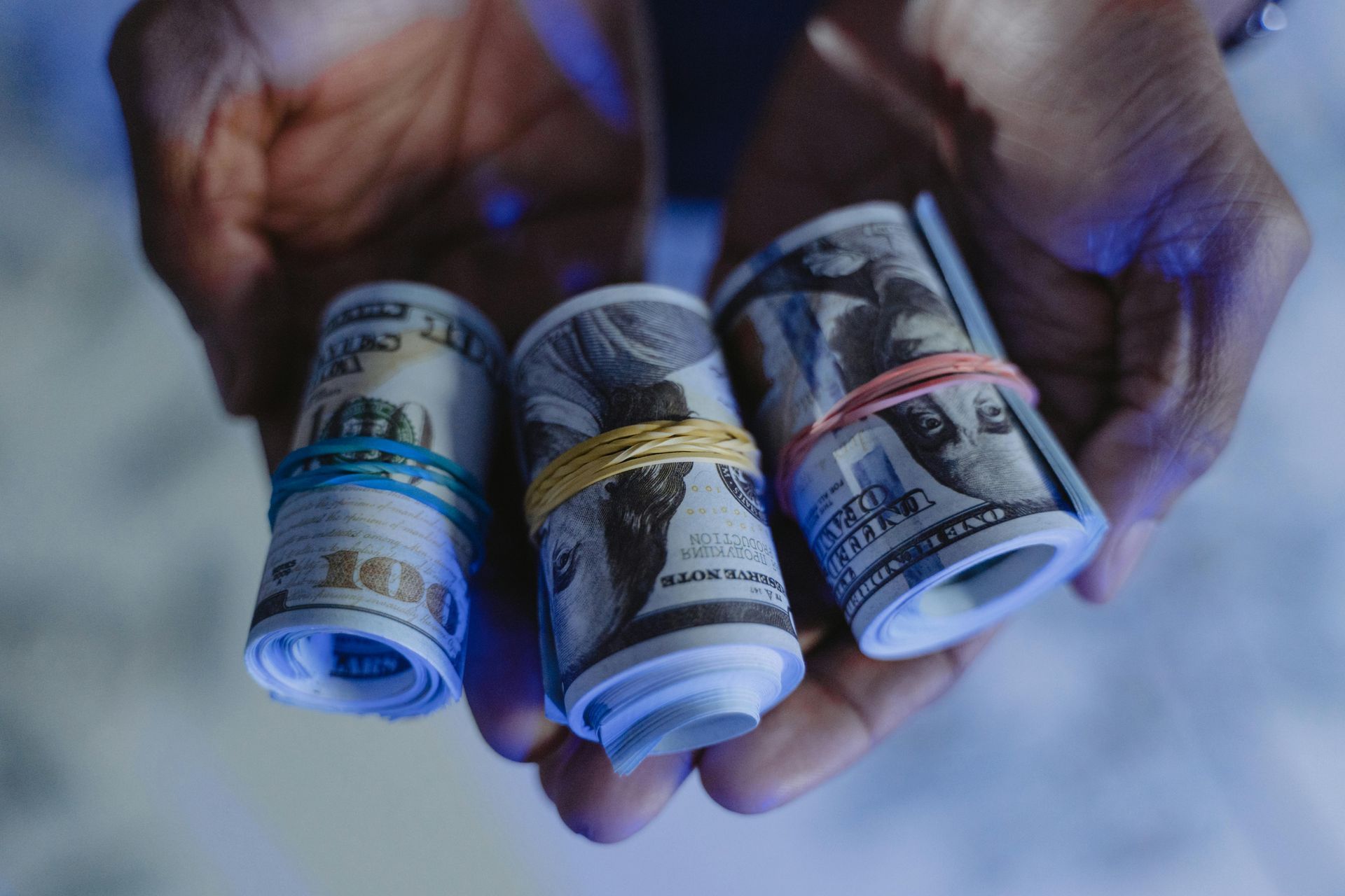 Hands holding three rolled-up bundles of US dollar bills secured with rubber bands.