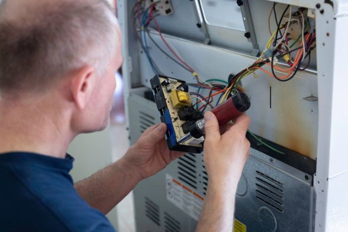 A man removing an electrical component from the back of a large appliance, he is holding it in his hands.