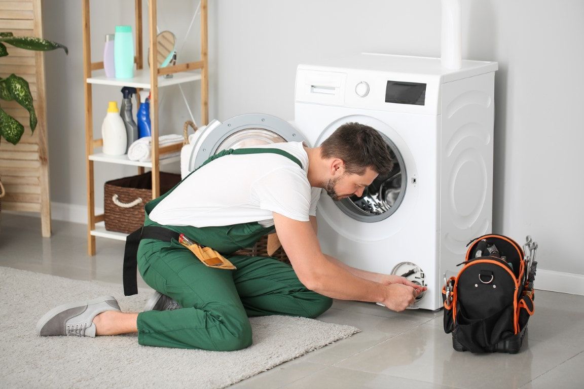 A man on his knees repairing a small section of a home dryer unit. He has a tool bag on the floor next to him.