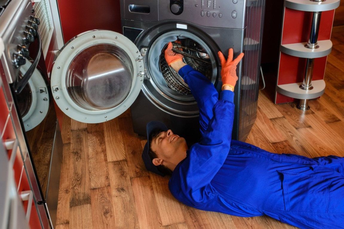 A man lying down on his back in a blue mechanic suit reaching up into a washing machine. He has orange gloves on.