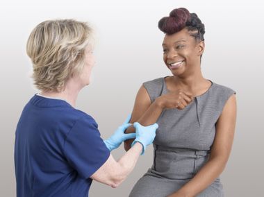 An older woman wearing medical gloves and a blue shirt is grasping the arm of a smiling younger woman in a gray dress.