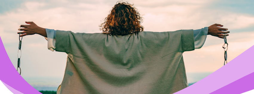 Person with outstretched arms standing outdoors at sunset, wearing a green jacket against a pink sky.