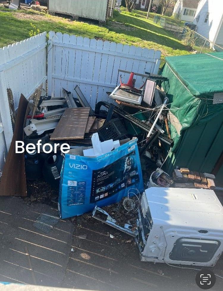 Cluttered backyard corner with broken furniture, boxes, and trash piled against white fences and a green tarp shed