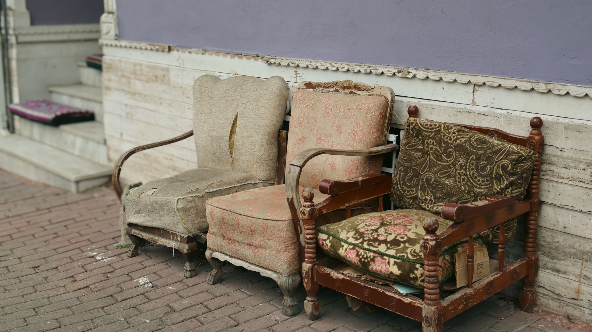 Three worn armchairs lined up on a brick sidewalk beside a white stone wall.