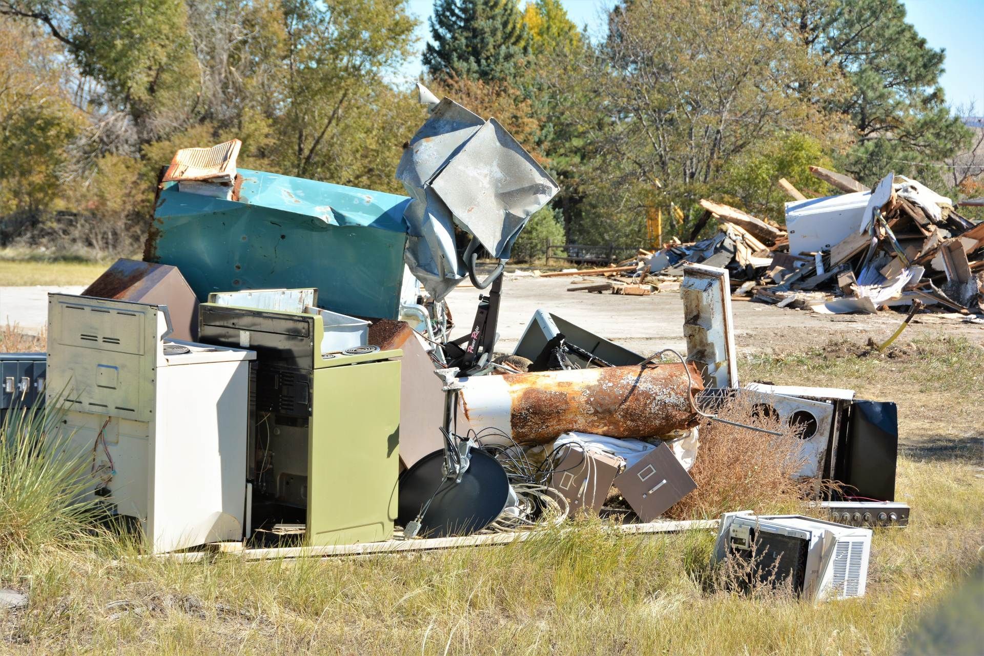 Piles of discarded appliances and debris scattered in a grassy outdoor area with trees in the background