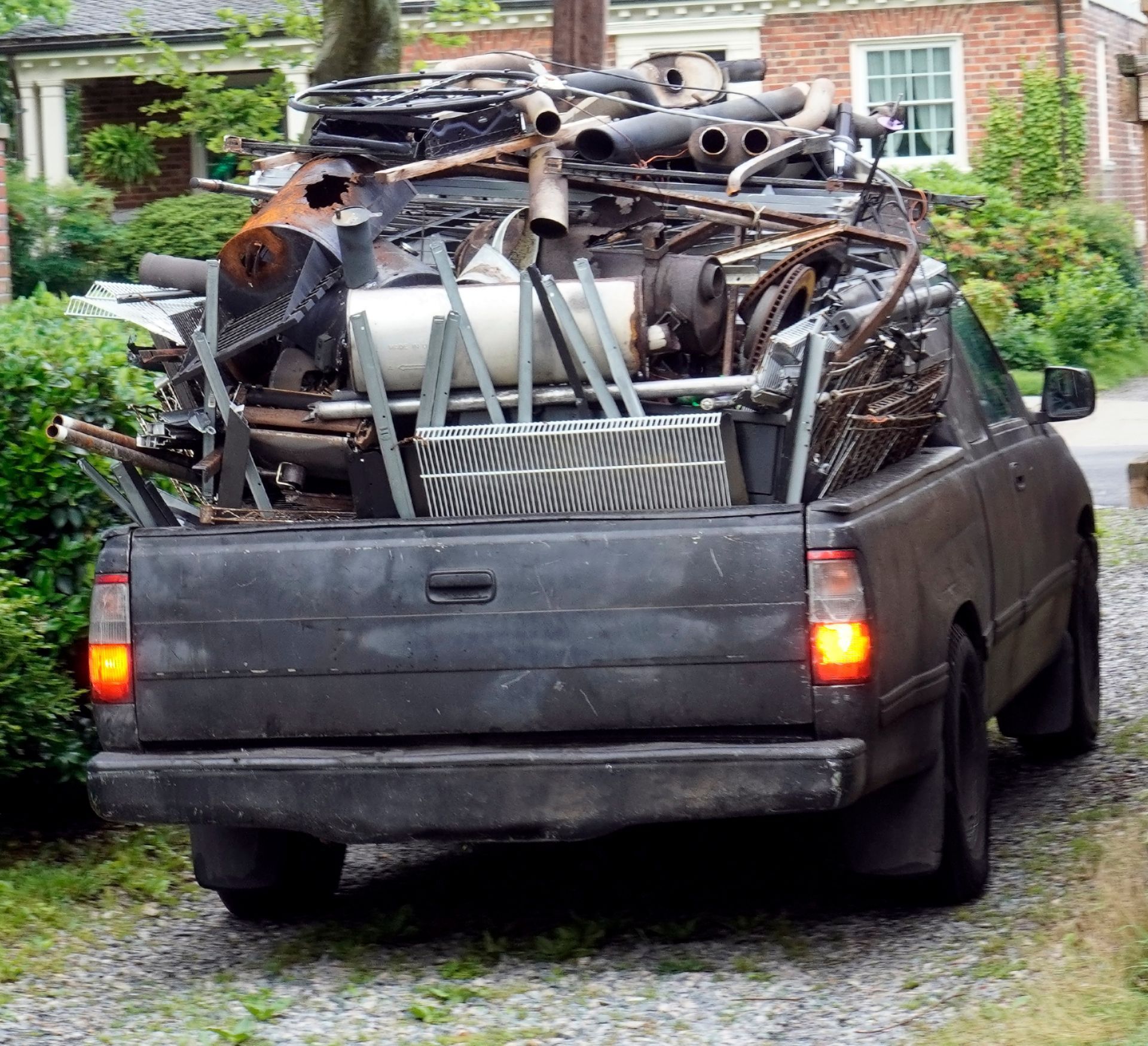 Black pickup truck overloaded with scrap metal and pipes in a gravel driveway near a brick house.