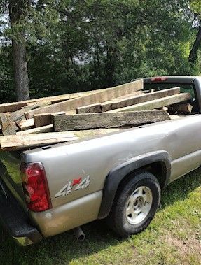 Silver pickup truck loaded with long wooden beams in the bed, parked on grass near trees.