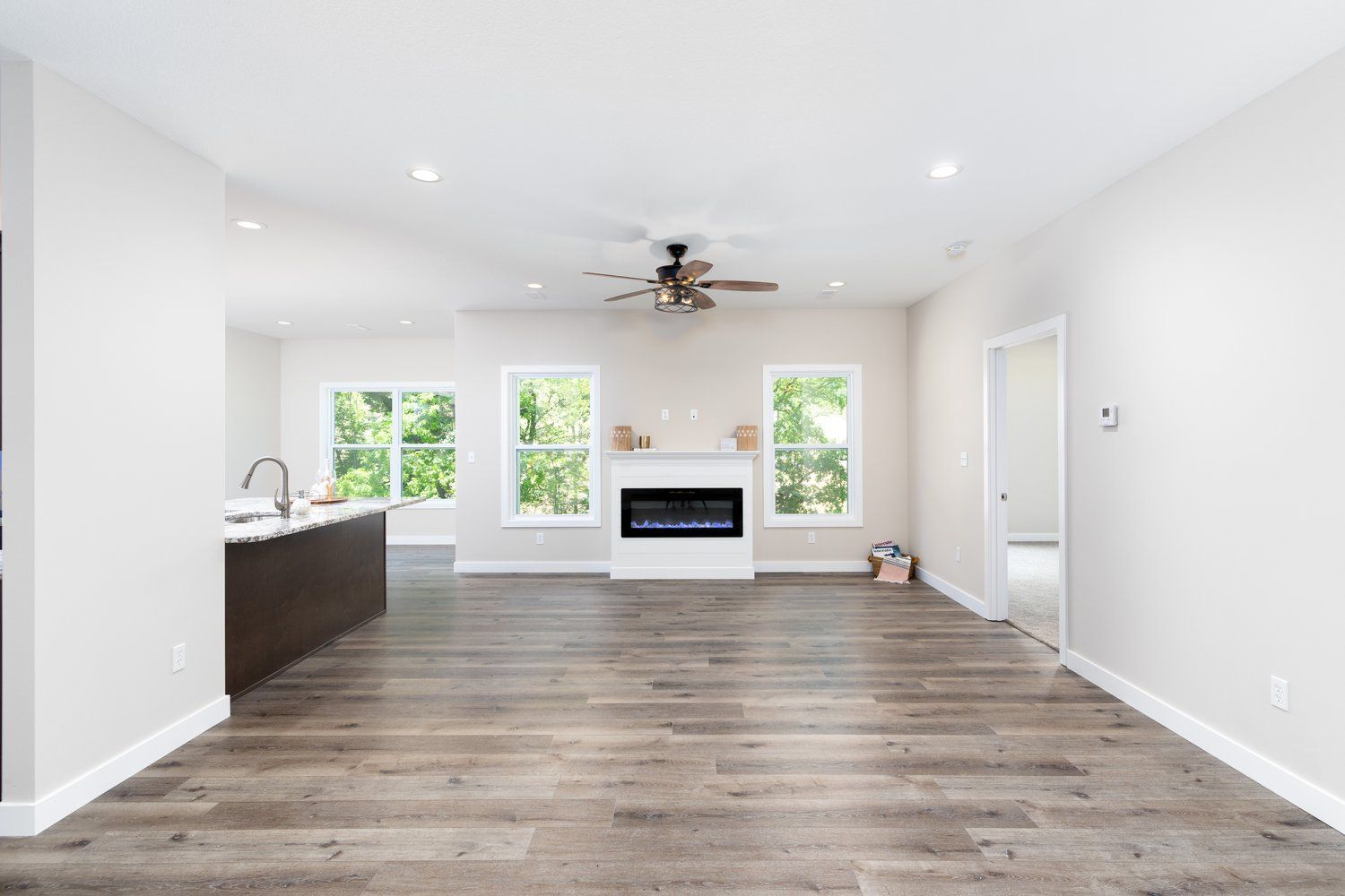 Empty modern living room with wood floors, white walls, ceiling fan, fireplace, and large windows.
