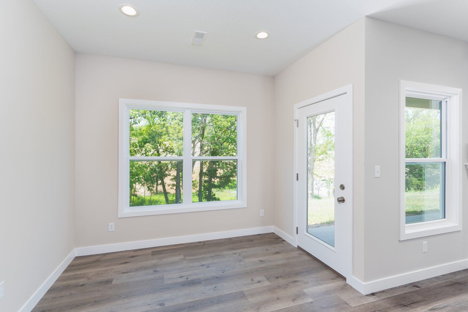 Bright empty room with white walls, gray wood floor, and large windows letting in natural light.