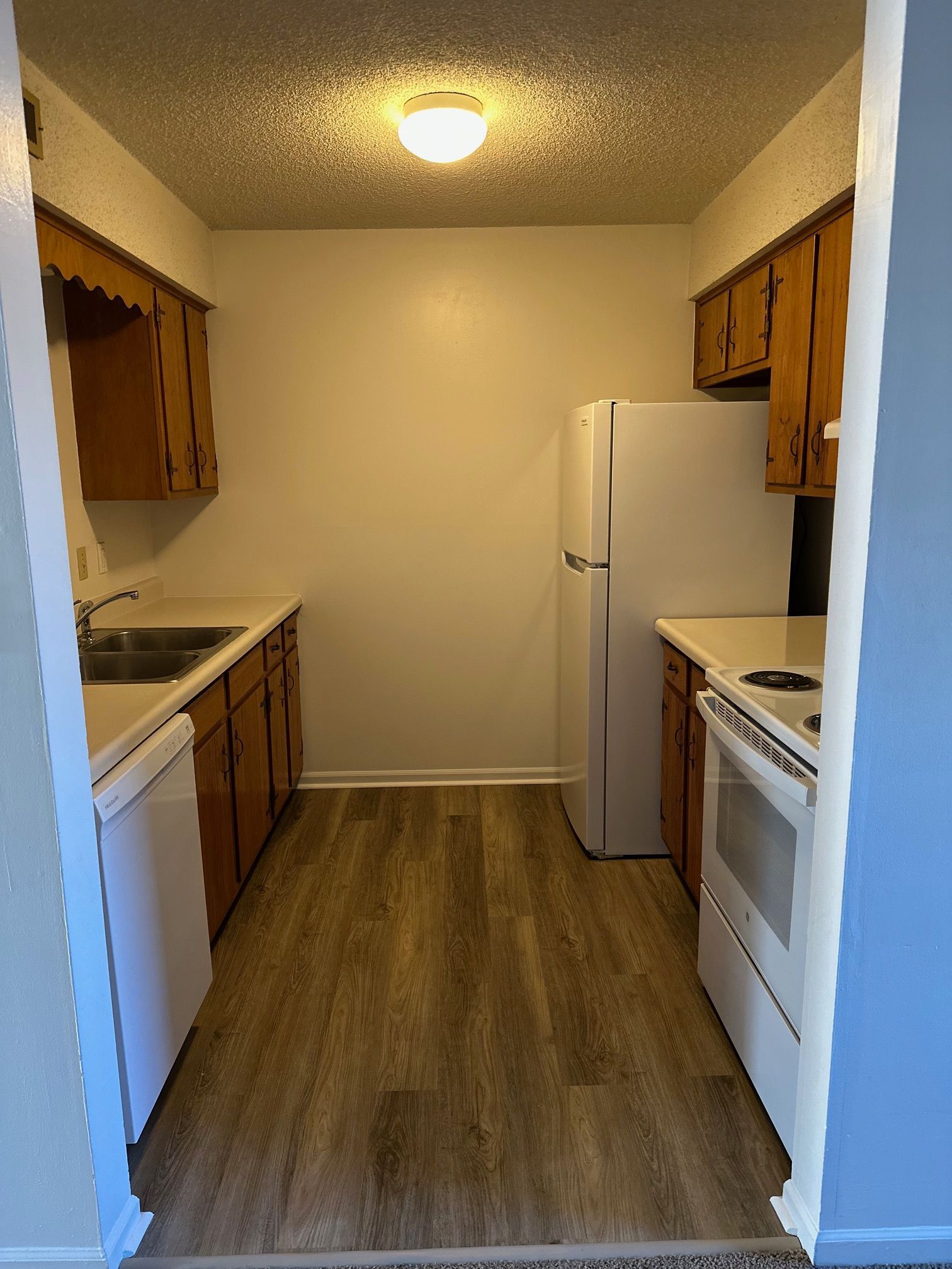 Narrow galley kitchen with wood cabinets, white appliances, and wood-look flooring under a ceiling light