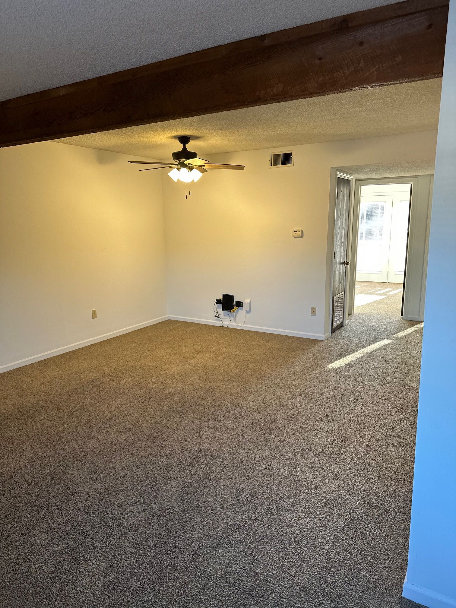Empty carpeted living room with beige walls, ceiling fan, exposed beam, and doorway to a hall