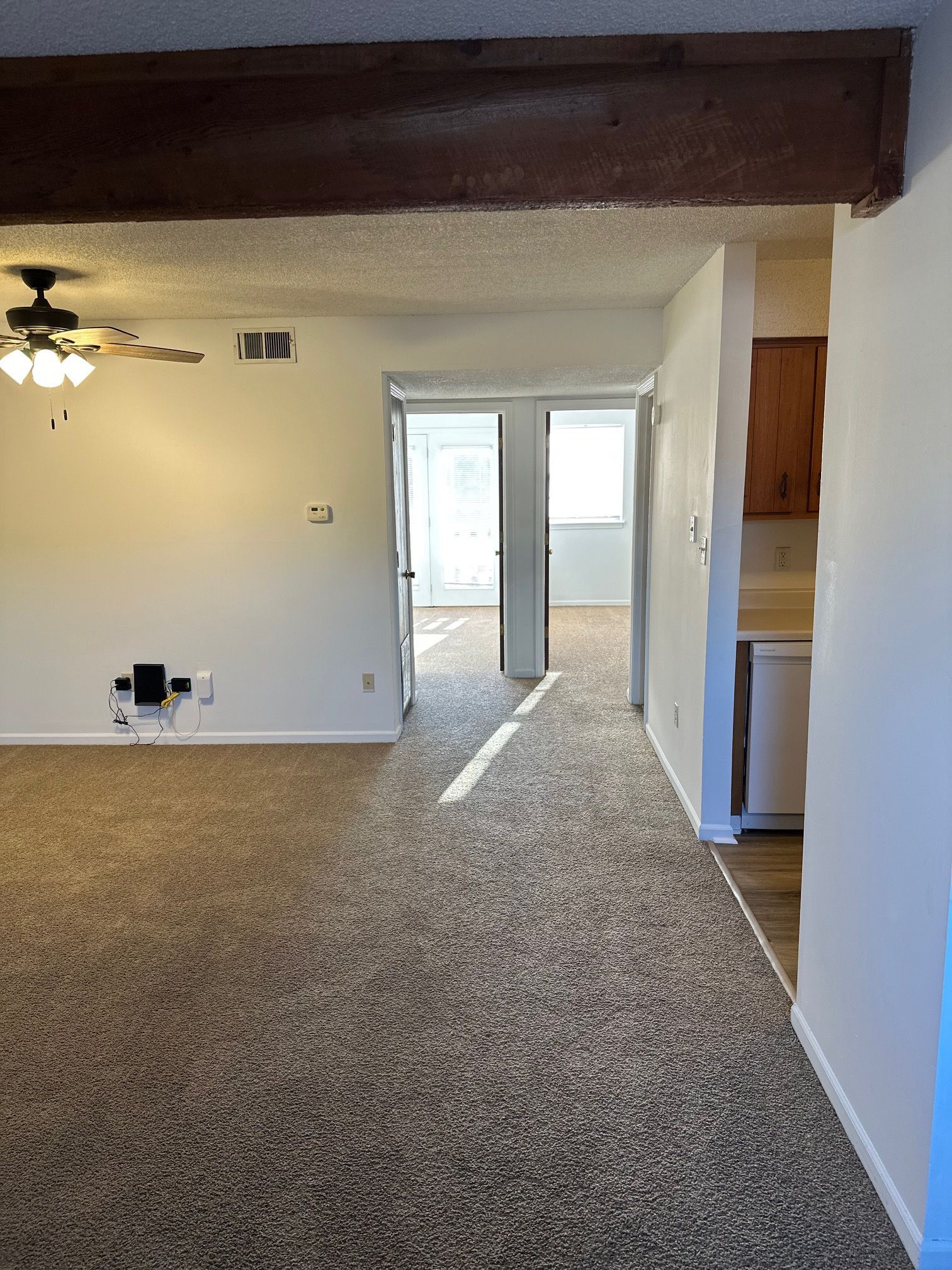Carpeted living room with exposed beam, ceiling fan, and open doorway to a bright adjoining room and kitchen.