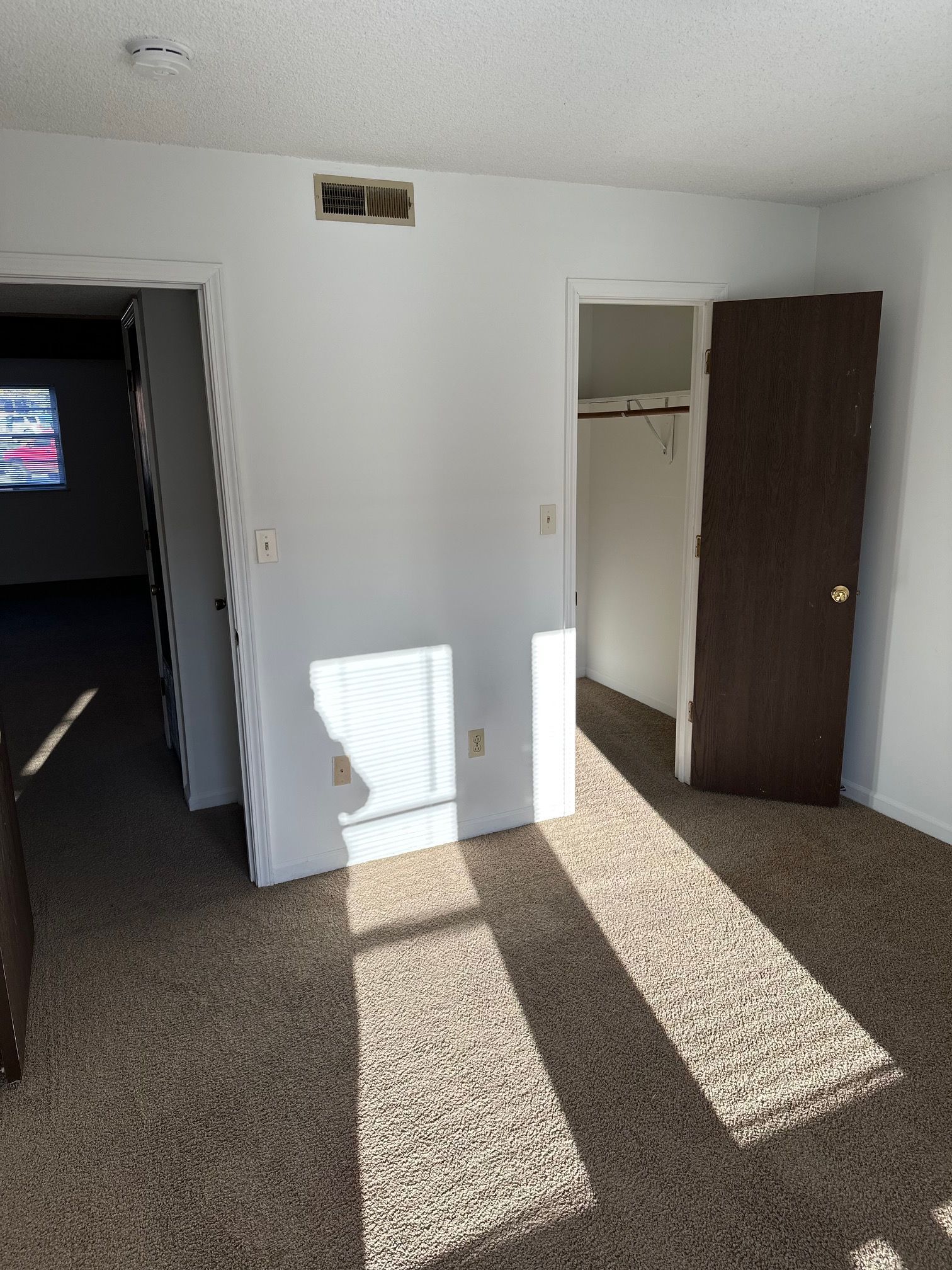 Empty sunlit room with open closet door and dark doorway, sunlight casting long shadows on carpet.