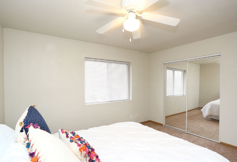 Bright bedroom with white bed, ceiling fan, window blinds, and mirrored closet doors