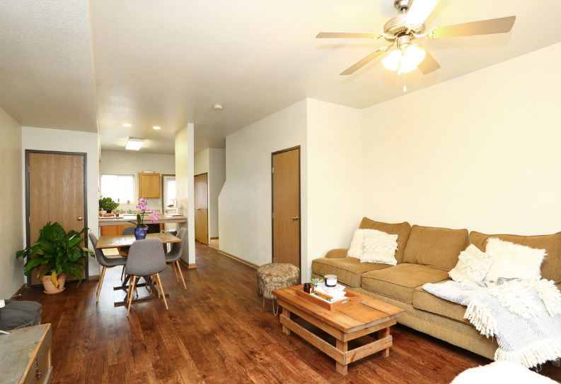 Bright living room with tan sofa, wooden floor, ceiling fan, and small dining area in back