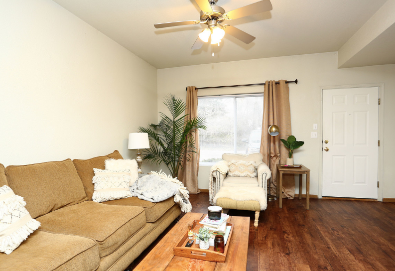 Bright living room with tan sofa, white chaise, coffee table, plants, and ceiling fan.
