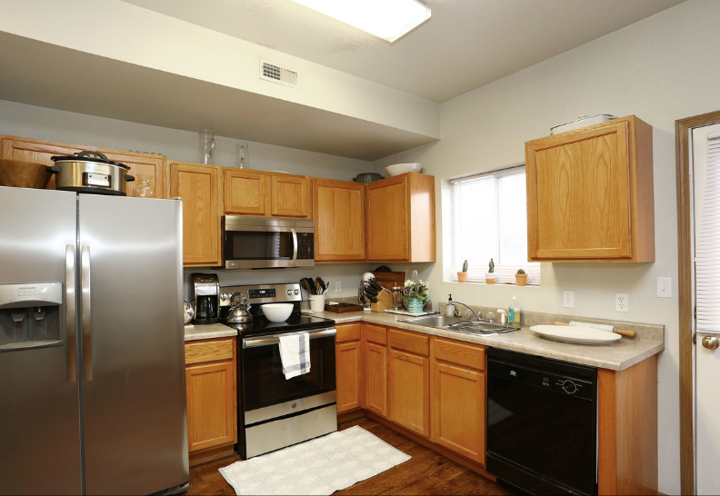 Bright kitchen with stainless steel fridge, black stove and dishwasher, wooden cabinets, and a window above the sink