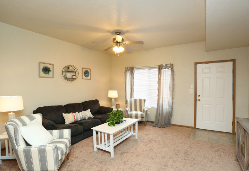 Cozy living room with dark sofa, white chair, coffee table, lamps, ceiling fan, and front door by window.