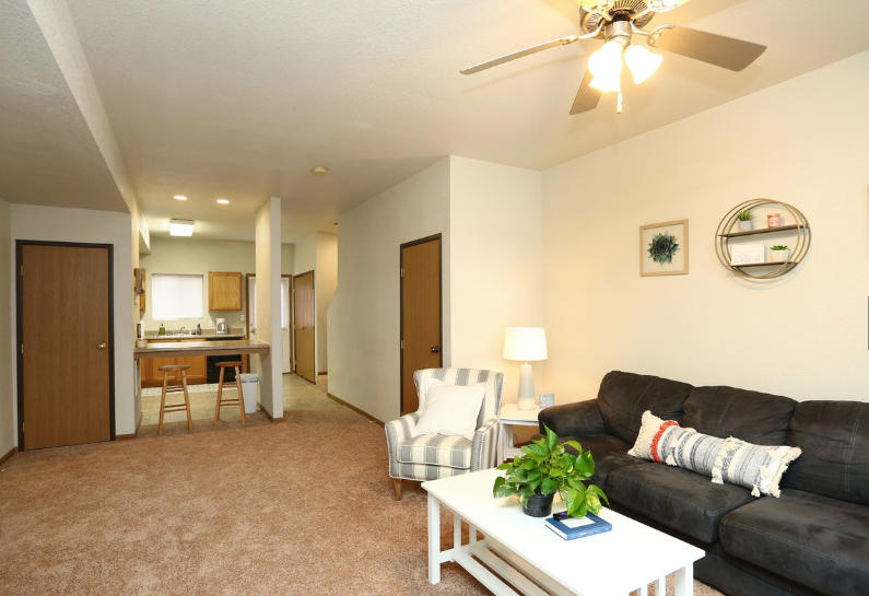 Cozy living room with dark sofa, white coffee table, ceiling fan, and open view to kitchen and hallway