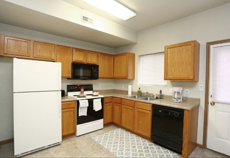 Empty kitchen with white appliances, wood cabinets, and a black dishwasher beside a window and door.