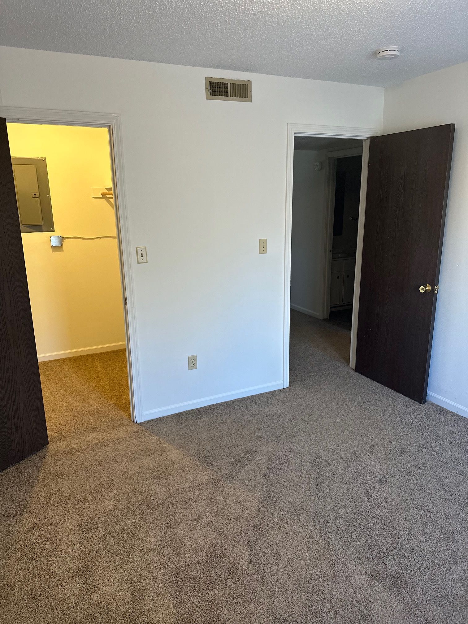 Empty bedroom with beige carpet, white walls, dark closet doors, and an open doorway to a lit hall