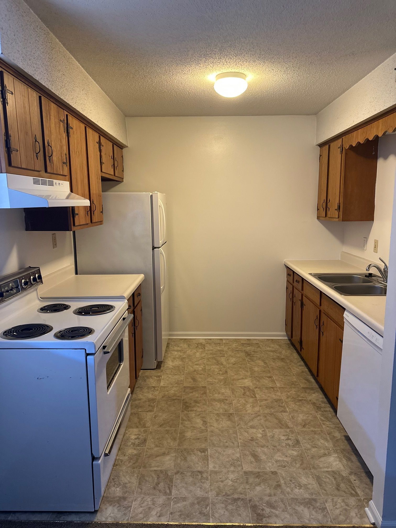 Small empty kitchen with wood cabinets, white appliances, and linoleum floor.