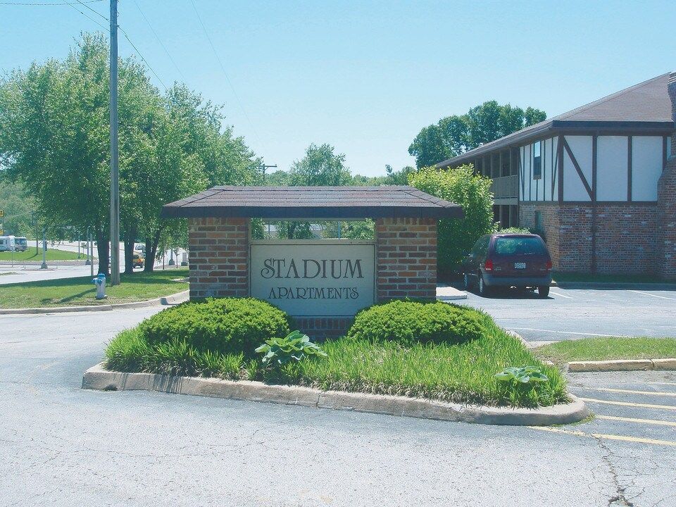 Brick stadium entrance sign with landscaped shrubs and a building in the background