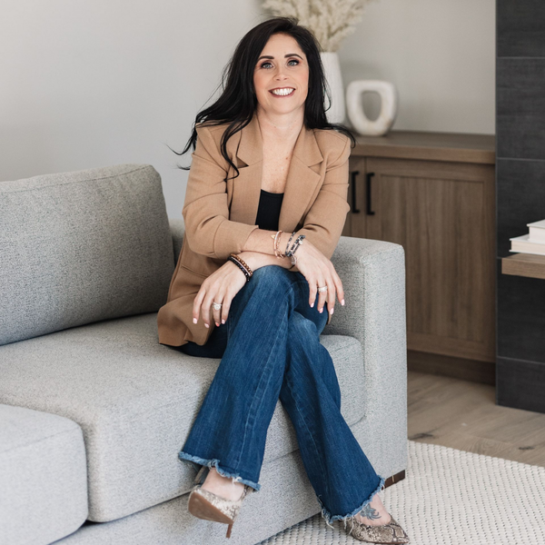 Woman seated on a gray sofa in a tan blazer and jeans, smiling in a modern living room.