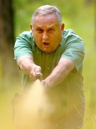 Man in green shirt, arms outstretched, pointing a gun with smoke.