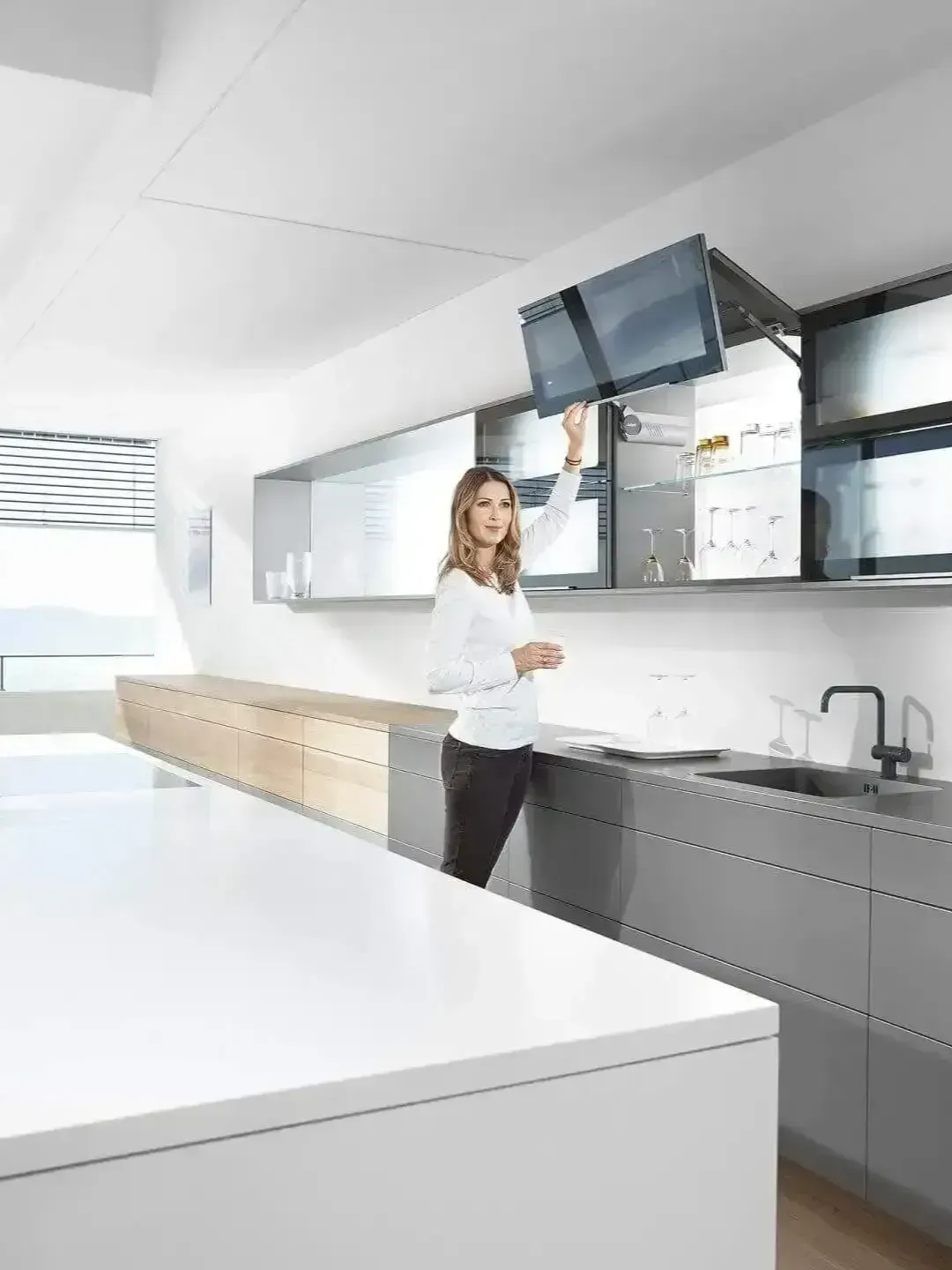 Bright modern kitchen with a woman standing at the counter near cabinets and a wall-mounted TV.