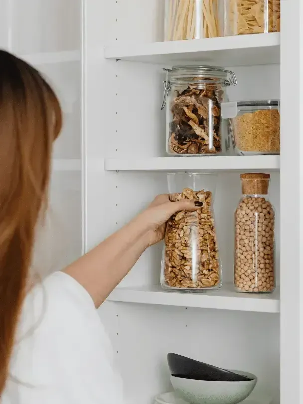Hand placing a jar of granola on a white pantry shelf with other storage jars.