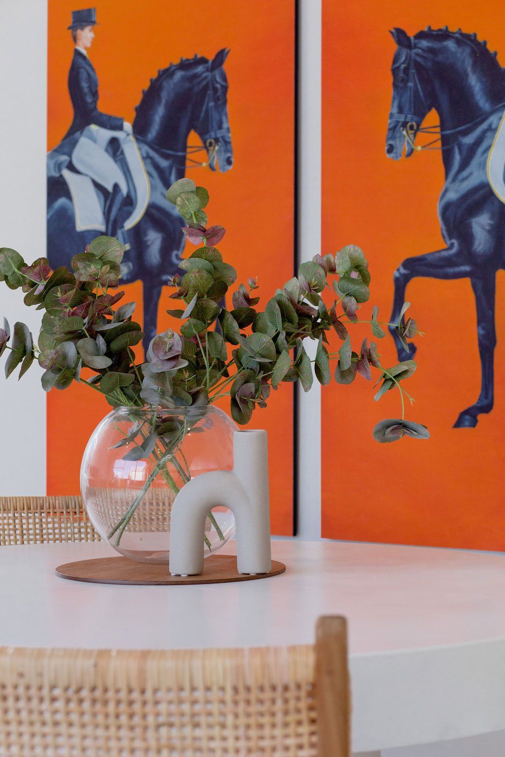 A vase with flowers in it is on a table in front of two paintings of people riding horses.